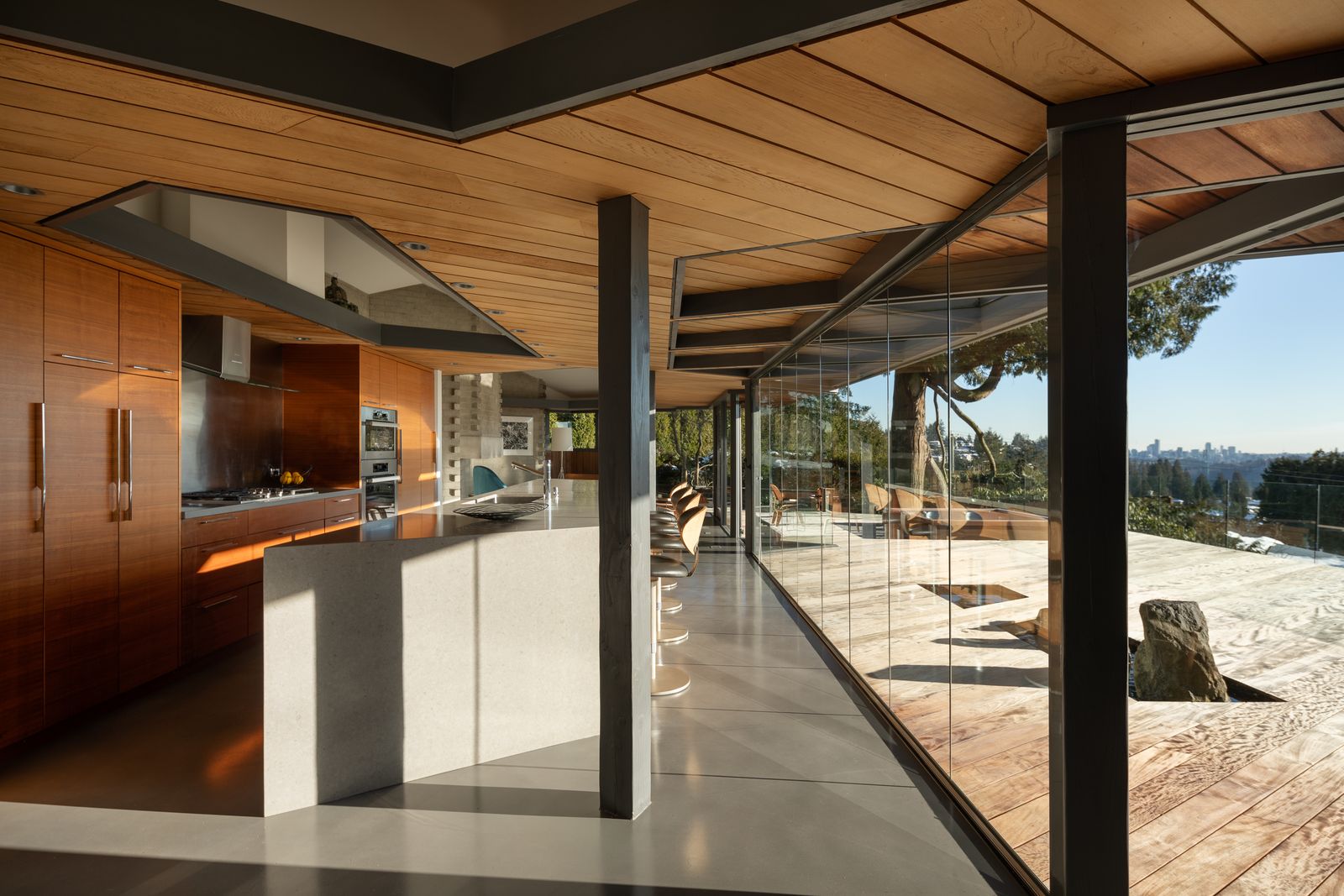 Huge kitchen with wooden ceiling and cupboards facing large windows with wooden terrace with a view.