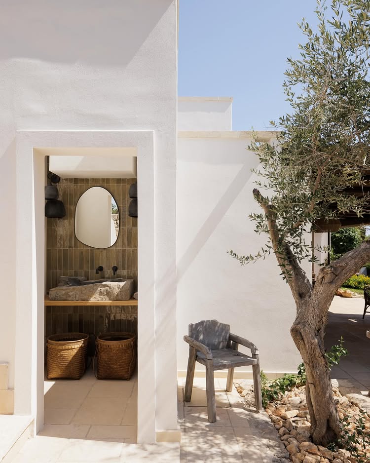 White walls of a masseria in Puglia, with a wooden chair, outside next to an olive tree and an open door showing a stone sink.